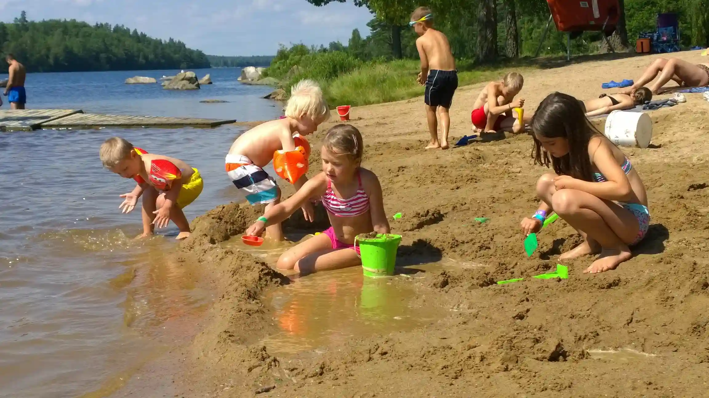 En grupp människor, inklusive barn, njuter av en solig dag på stranden vid Långasjönäs camping & stugby. De flesta bär badkläder och har kul vid stranden under en sommardag.