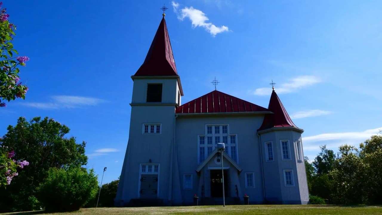 Bild av en vit träkyrka med spetsigt torn och kors, omgiven av grönska under en klarblå himmel vid Fällfors Camping.