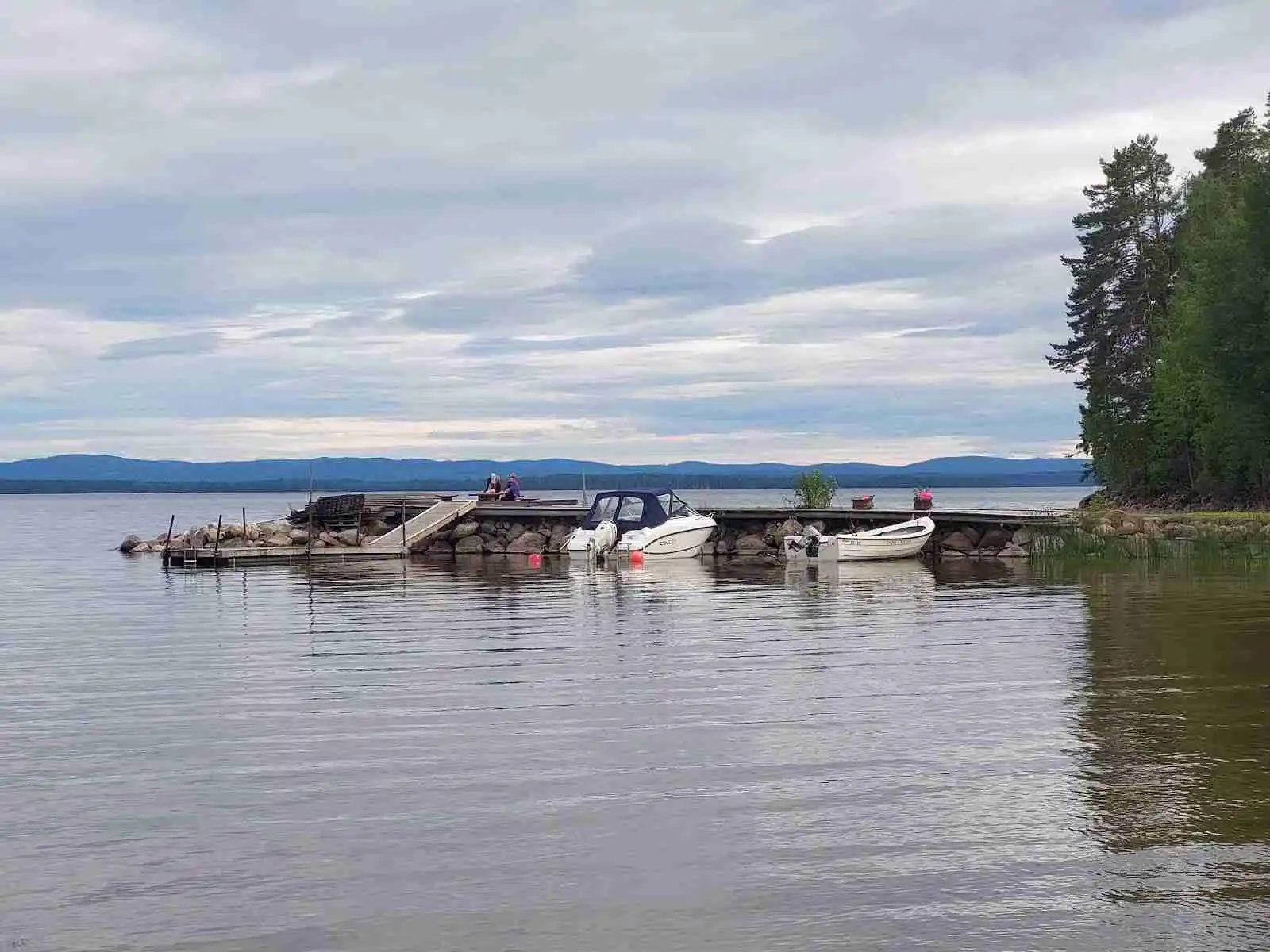 En motorbåt ligger förtöjd vid en brygga på en sjö med omgivande gröna kuster och molnig himmel, vid Norra Dellen Familjecamping.