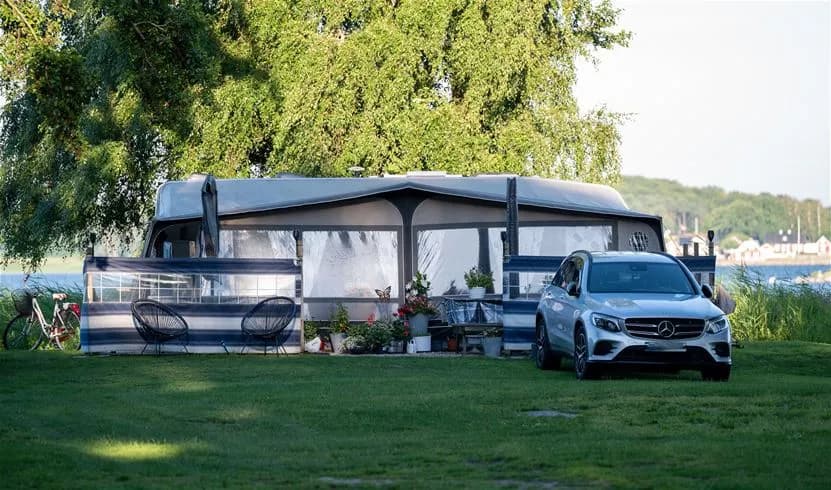 En husvagn med uppfälld markis står parkerad på en campingplats. En bildörr är öppen, vilket ger en inbjudande känsla på den gröna omgivningen vid My Camping Tredenborg.