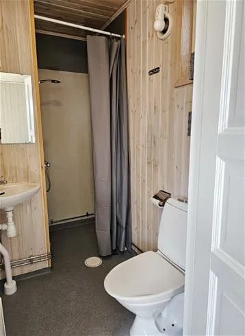 En campingtoalett med toalettstol, handfat och duschdraperi. Ljust kaklat golv och en enkel, funktionell inredning.