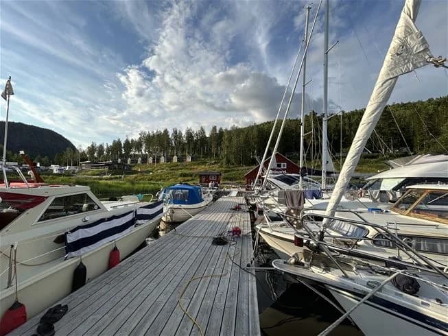 En småbåtshamn vid Skuleberget Havscamping där flera båtar ligger förtöjda vid bryggan, omgivna av vacker natur och spegelblankt vatten. Skogsklädda berg reser sig i bakgrunden.