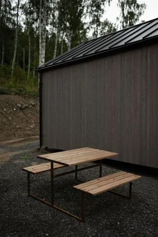 En träbänk och ett bord i trä vid en campingplats omgivna av natur, med en enkel träkonstruktion i bakgrunden.