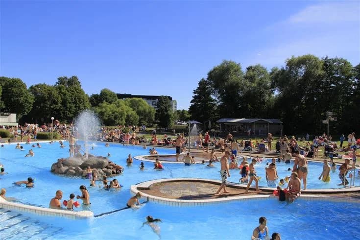 En grupp människor njuter av en solig dag vid en stor utomhuspool. Vattenrutschkanor och badande barn syns i bakgrunden, omgivna av gröna träd och sommarhimmel. Campingen Fyrishov erbjuder avkoppling och nöje i natursköna omgivningar.