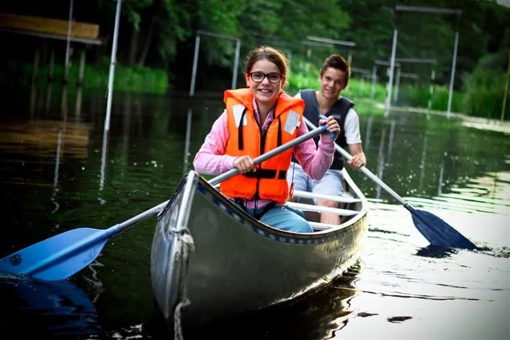 Person paddlar i en kanot på en lugn vattenström omgiven av grönska, iklädd flytväst och leende.