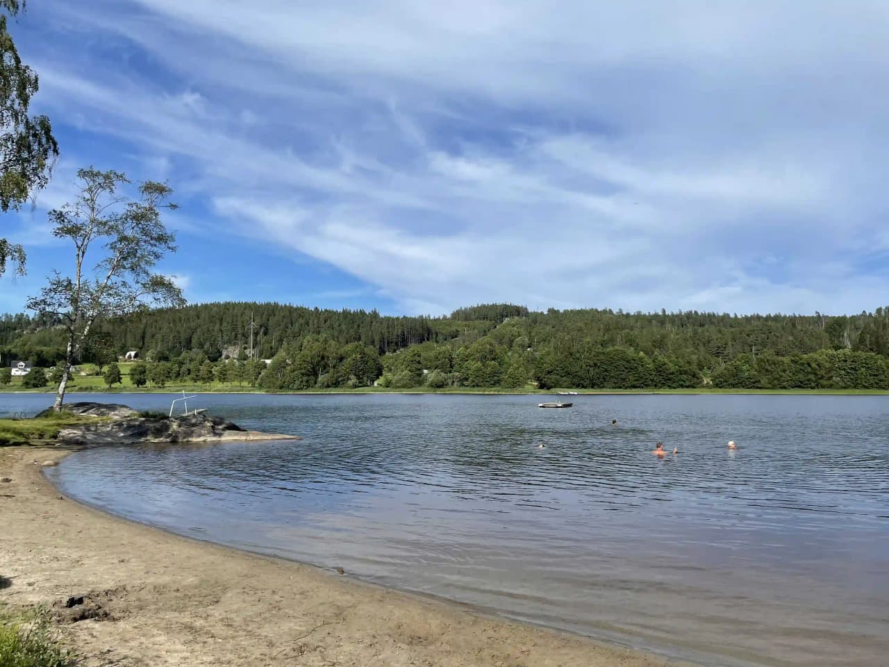 En somrig dag vid sjökanten på Bullarebygdens Familjecamping, med en stilla vattenyta och lummiga stränder runt omkring.