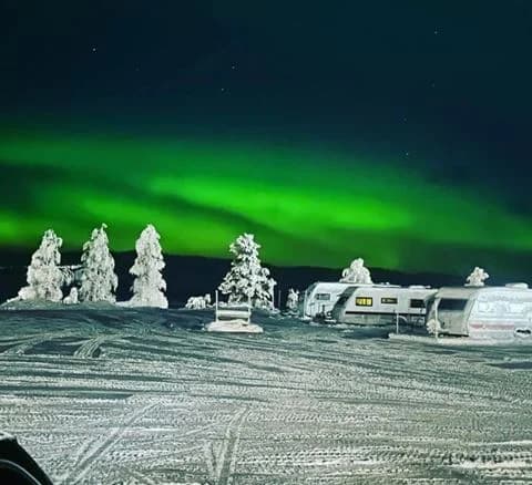 En snötäckt campingplats vid midnatt med en husbil och lastbil parkerade. Norrskenet lyser upp den mörka vinternatten.