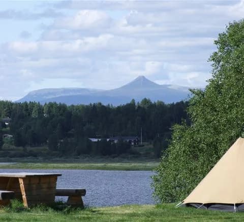 En campingplats i Särna med ett tält uppsatt på en gräsbevuxen äng vid en reservoar. Området omges av vidsträckta gräsmarker och en naturskön miljö.