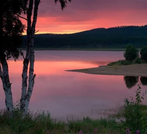 En vacker bild av Särna Camping vid solnedgången med ett stilla vattendrag i förgrunden och horisonten synlig i fjärran, omgiven av naturlandskap.