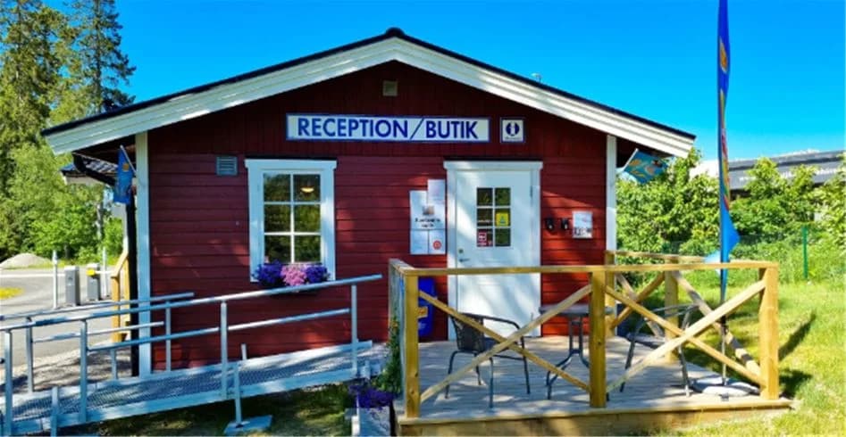 En mysig stuga omgiven av grönska på Trollhättans Camping City. Stugan har en träfasad, en liten veranda, och står på en välskött tomt med skugga från omgivande träd.