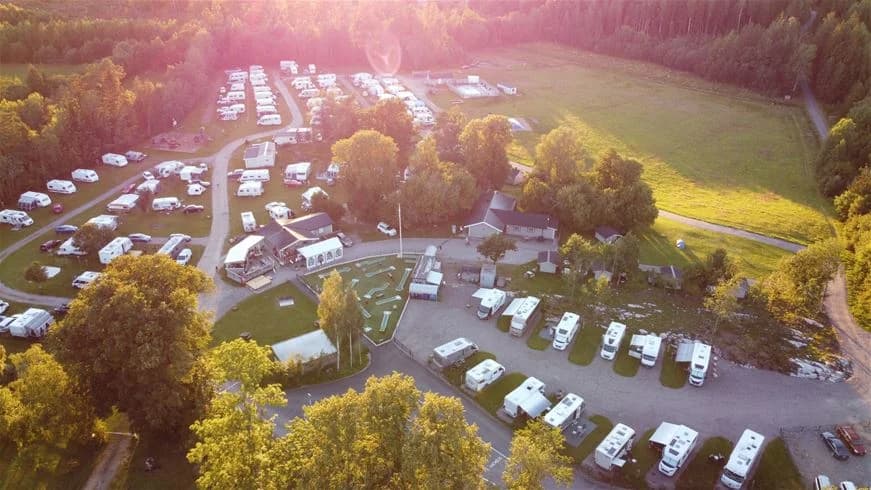 Fågelperspektiv över Stenrösets Camping i Trollhättan, med husbilar, tält och husvagnar uppställda på en asfalterad parkeringsplats omgiven av grönska.
