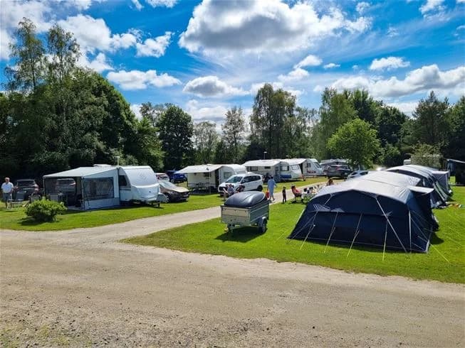 En livlig campingplats i Trollhättan med tält, husvagnar och husbilar uppställda. I bakgrunden syns en parkering och moln på himlen.