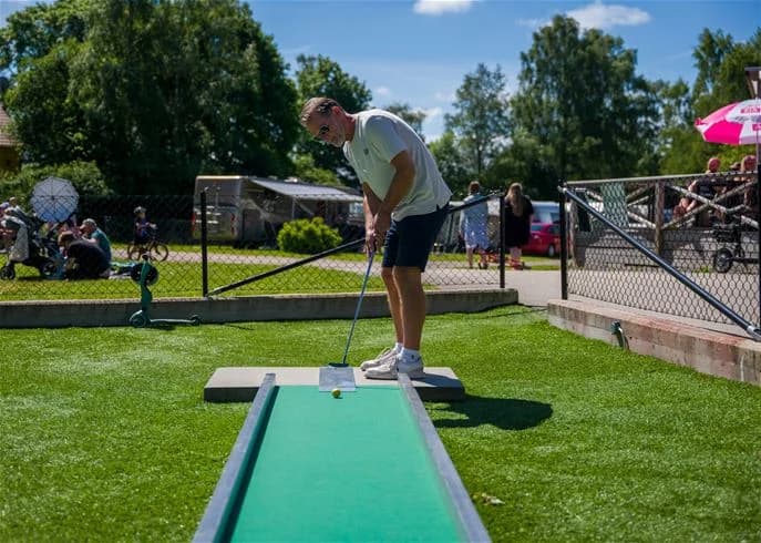 Personer som spelar minigolf på en grönskande camping. Soligt väder och glada människor ger en känsla av fritid och nöje.