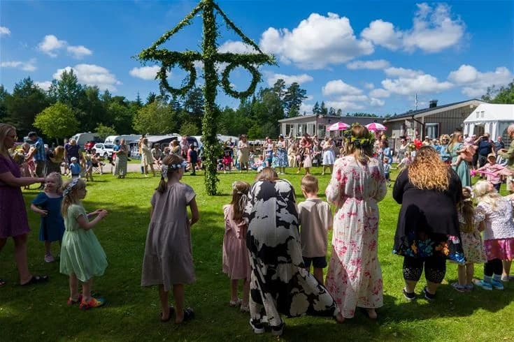 En grupp människor deltar i ett evenemang på Stenrösets camping i Trollhättan. De är klädda i somriga kläder och njuter av fritidsaktiviteter. I bakgrunden syns en stång, vilket kan tyda på en traditionell festlighet.