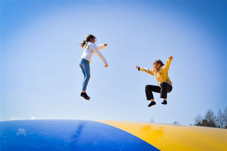 En grupp glada människor hoppar i luften vid Stenrösets camping i Trollhättan, omgivna av natur och blå himmel, vilket skapar en känsla av fritid och semester.