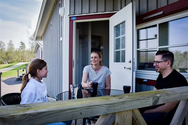 En glad familj sitter och njuter av solen vid sitt campingområde, med stolar och en grön gräsmatta som omger dem. En husvagn syns i bakgrunden, tecken på en härlig campingupplevelse på Stenrösets Camping i Trollhättan.
