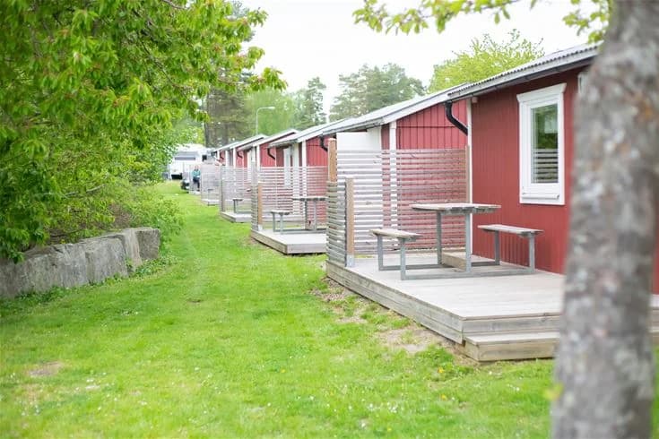En mysig stuga omgiven av grönska vid Kinnekulle camping & stugby. Stugan har en liten veranda och ligger i en välskött trädgård med buskar och en gräsmatta som skapar en inbjudande atmosfär.