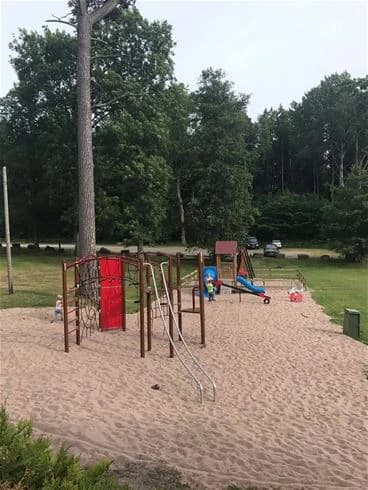 En lekplats på Kinnekulle Camping & Stugby med gungor, rutschkana och andra lekredskap, omgiven av ett grönskande parkområde.