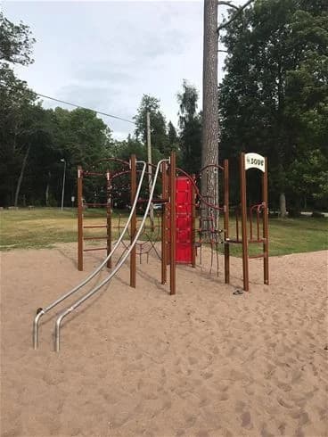 Bild av Kinnekulle Camping & Stugby som visar en livlig lekplats med rutschbana, omgiven av grönskande natur. Lekutrustning är placerad på en öppen yta, vilket skapar en inbjudande miljö för barn att leka i.