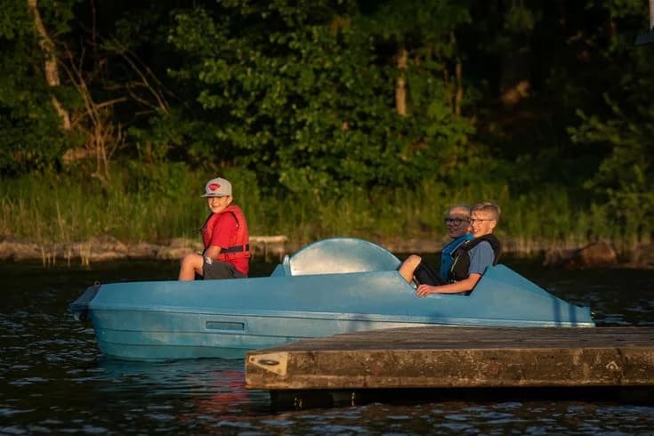En motorbåt glider fram på en stilla sjö vid Kinnekulle camping & stugby, omgivna av frodig grönska och blå himmel, vilket skapar en idyllisk och avkopplande miljö för rekreation och friluftsliv.