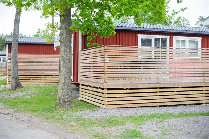 En mysig trästuga med träpanel och altan på Kinnekulle camping & stugby, omgiven av ett staket och grönska i skuggan.