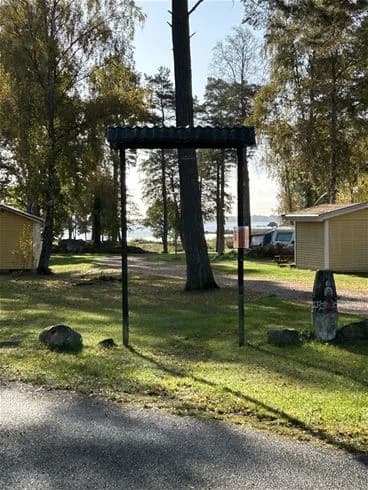 En grönskande campingplats med en stor gräsmatta, några trädstammar och skuggade områden. En skyltstolpe syns i bakgrunden.