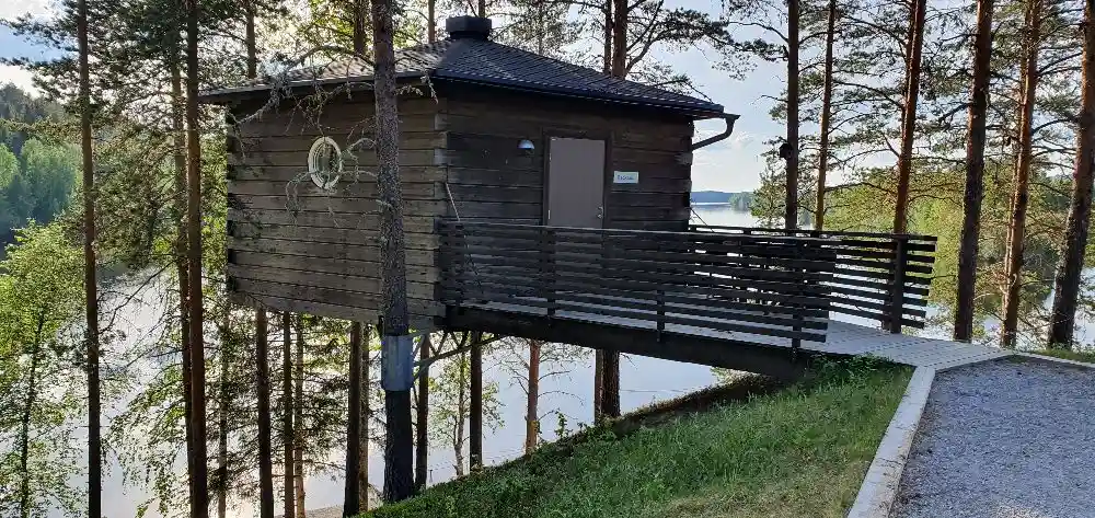 Bild av en trästig med räcken som leder upp till en timmerstuga, omgiven av tät skog.