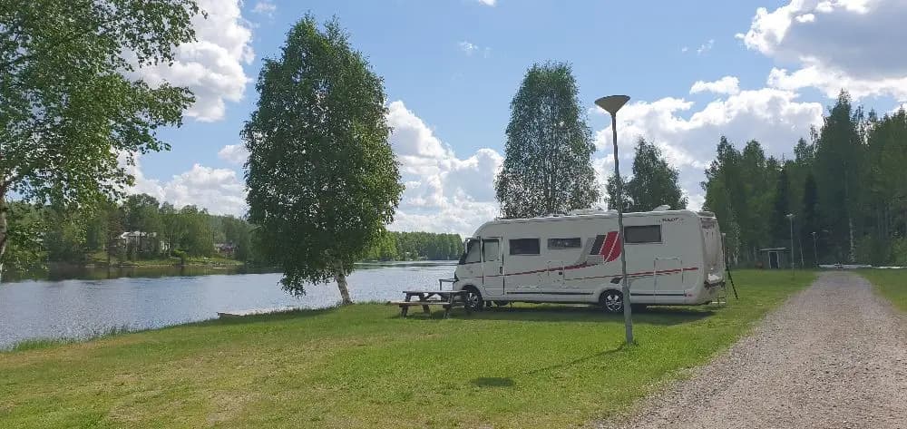 En naturskön campingplats vid Granö Beckasin med husbilar och husvagnar uppställda på en grön gräsyta. Omgiven av träd och en liten damm i bakgrunden.