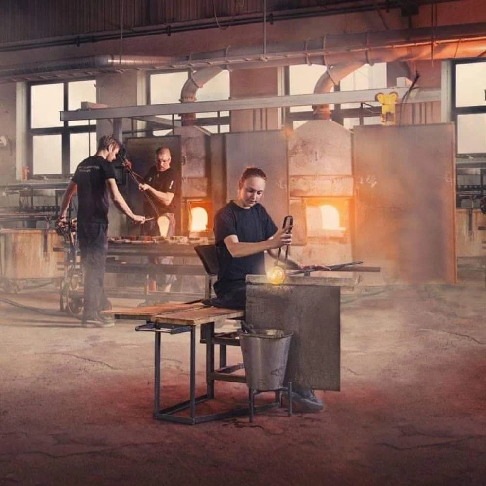 En glasarbetare som jobbar i verkstaden på Målerås glasbruk, omgiven av eld och traditionella verktyg i en industriell miljö.