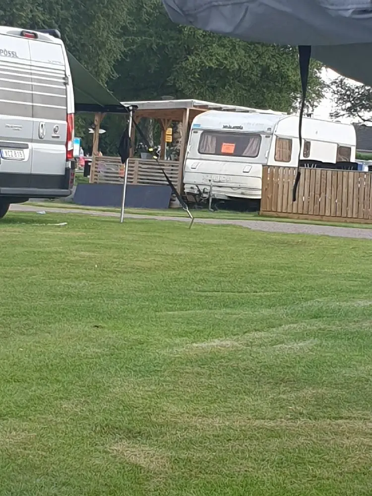 Alt-text: "Vy över Öjaby Camping med husvagn, grön gräsmatta och skuggiga träd i bakgrunden, samt en parkerad skåpbil i närheten."