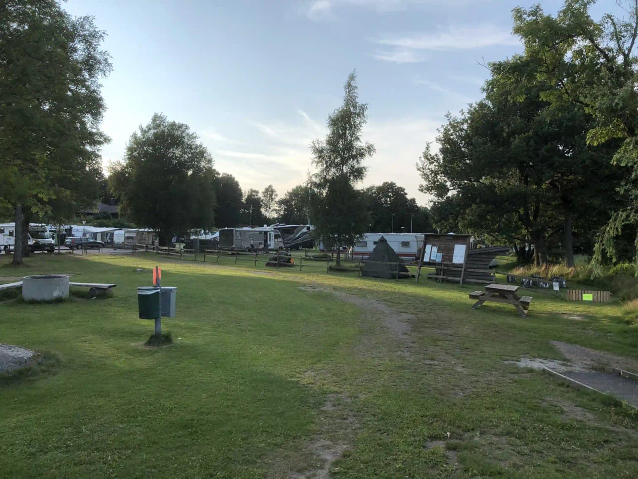 En grönskande campingplats med husvagnar och bilar i närheten av en gräsmatta och träd. Det finns även en utomhusbänk i parken.
