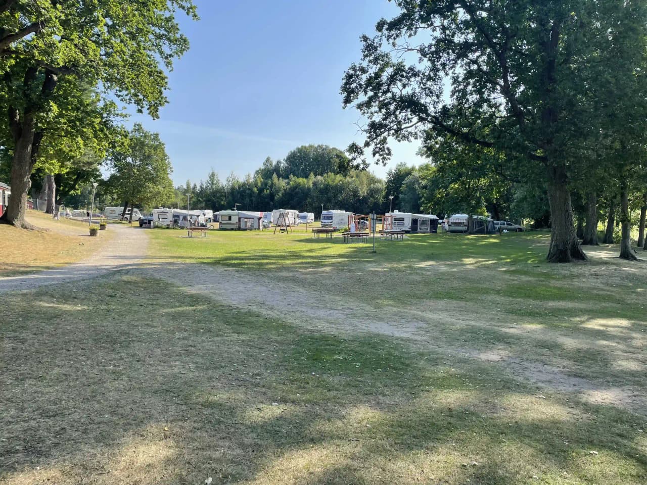 En grön campingplats med träd och gräs, där flera husvagnar är parkerade i skuggiga områden. Området ser lugnt och välskött ut, idealiskt för avkoppling.