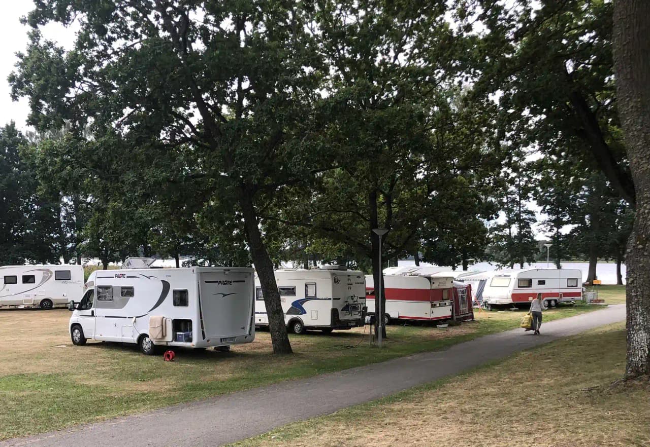 En husvagn parkerad i skuggan på en campingplats omgiven av träd och andra husvagnar, med en lastbil synlig i bakgrunden.