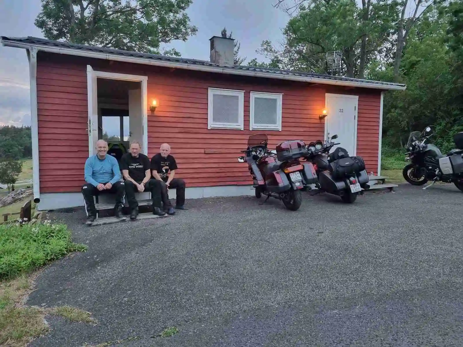 En idyllisk campingplats vid Vättersmålen med små stugor och husvagnar uppställda nära vattnet. En bil och en motorcykel står parkerade framför en stuga, omgiven av lugn natur.
