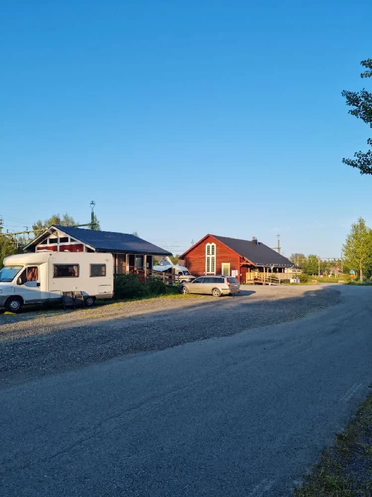 En vit byggnad med rött tak i en lantlig miljö, omgiven av asfaltväg och parkeringsplats.