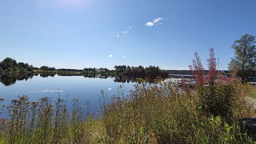 Bild av Trångsvikens bygdegård med en klarblå sommarhimmel i bakgrunden. Det finns en vattenyta i förgrunden som ger en harmonisk känsla av natur och lugn.