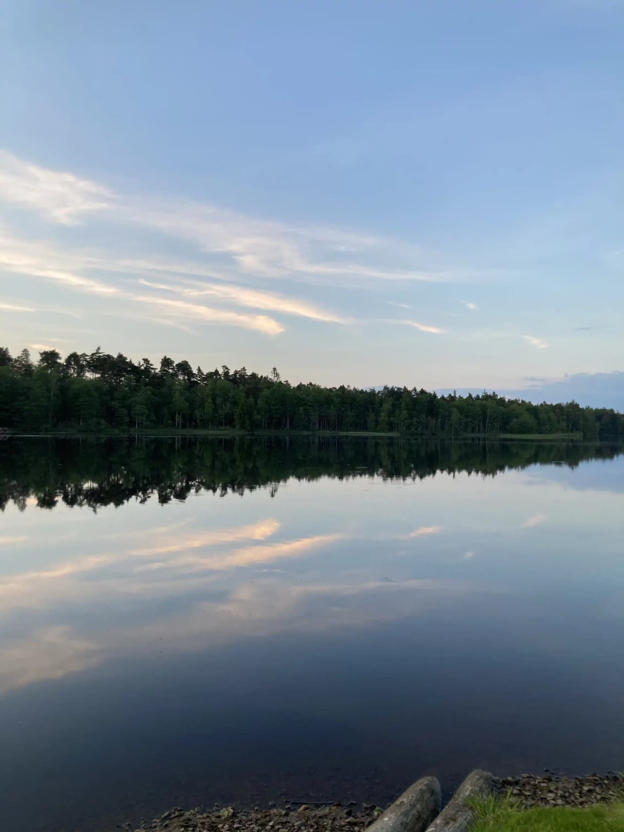 En stilla sjö med en spegelblank yta under en blå himmel med moln. I horisonten syns skogsområden som reflekteras i vattnet, vilket skapar en fridfull och naturskön vy.