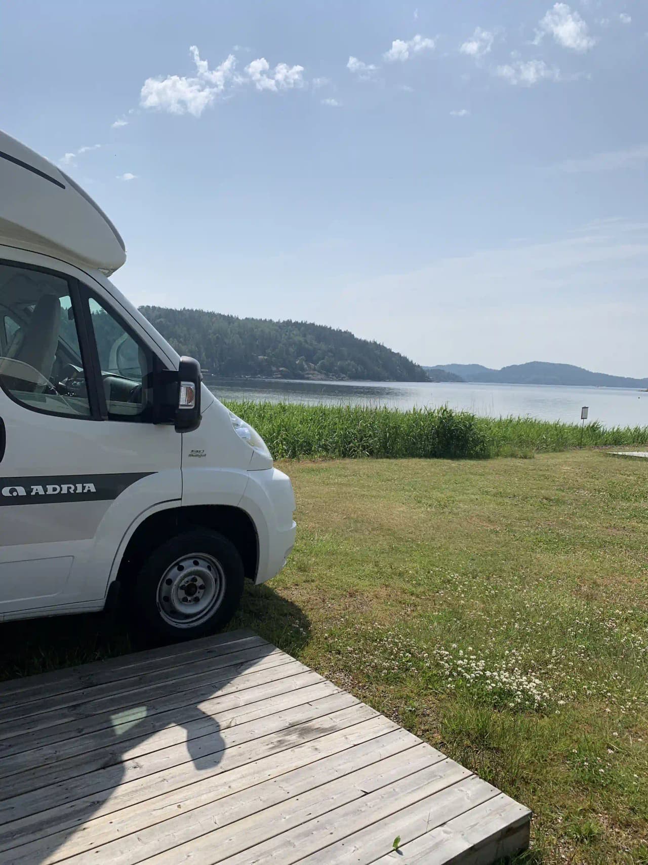 En husbil parkerad vid sidan av en grusväg nära skogsbryn, sett vid Vindöns Camping och Marina. I bakgrunden sträcker sig grönskande träd och en klar himmel.
