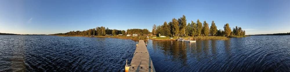 En pittoresk vy över Målsånna Camping Park med en stilla blå sjö och horisontlinje i bakgrunden. Sjön speglar omgivningen och en liten brygga sträcker sig ut över vattnet, vilket skapar en naturskön och avkopplande atmosfär.