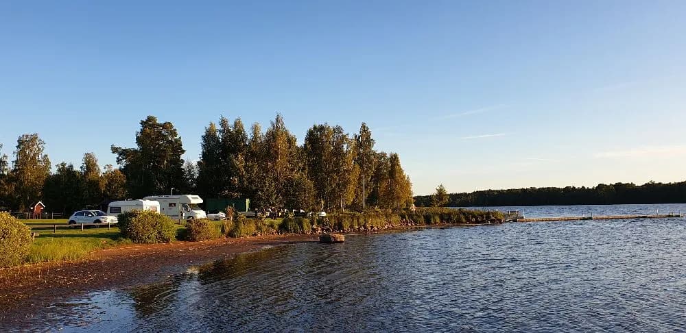 Bild av en lugn vattensamling vid Målsånna Camping Park, omgiven av skogsklädda stränder och en klar blå himmel.