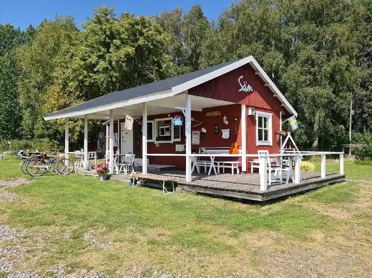 Bild av Säms Camping och Café: En inbjudande stuga med veranda, belägen på en välskött gräsmatta. Stugan har ett brunt tak och en vit dörr, omgiven av lugna utomhusmiljöer som skapar en trivsam atmosfär för besökarna.