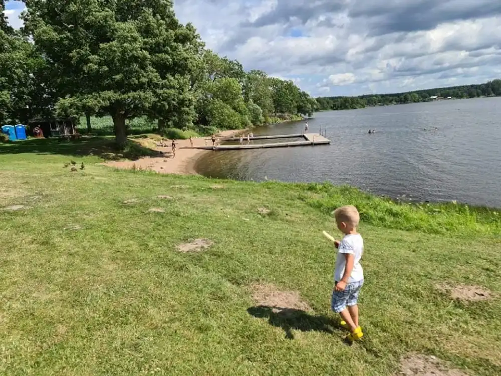 Vy över Säms camping och café, med en vacker sjö i förgrunden som kantas av grönska. Besökare njuter av sommarens härliga väder längs strandlinjen och deltar i olika fritidsaktiviteter.