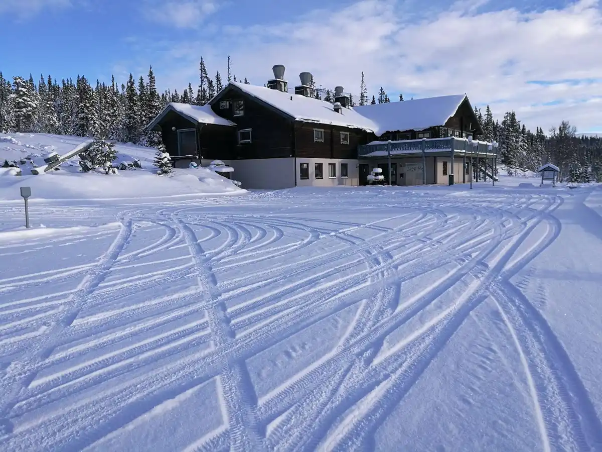 En snötäckt stuga vid Idre Fjäll Camping omgiven av vinterlandskap med skidbackar och larixträd i bakgrunden.