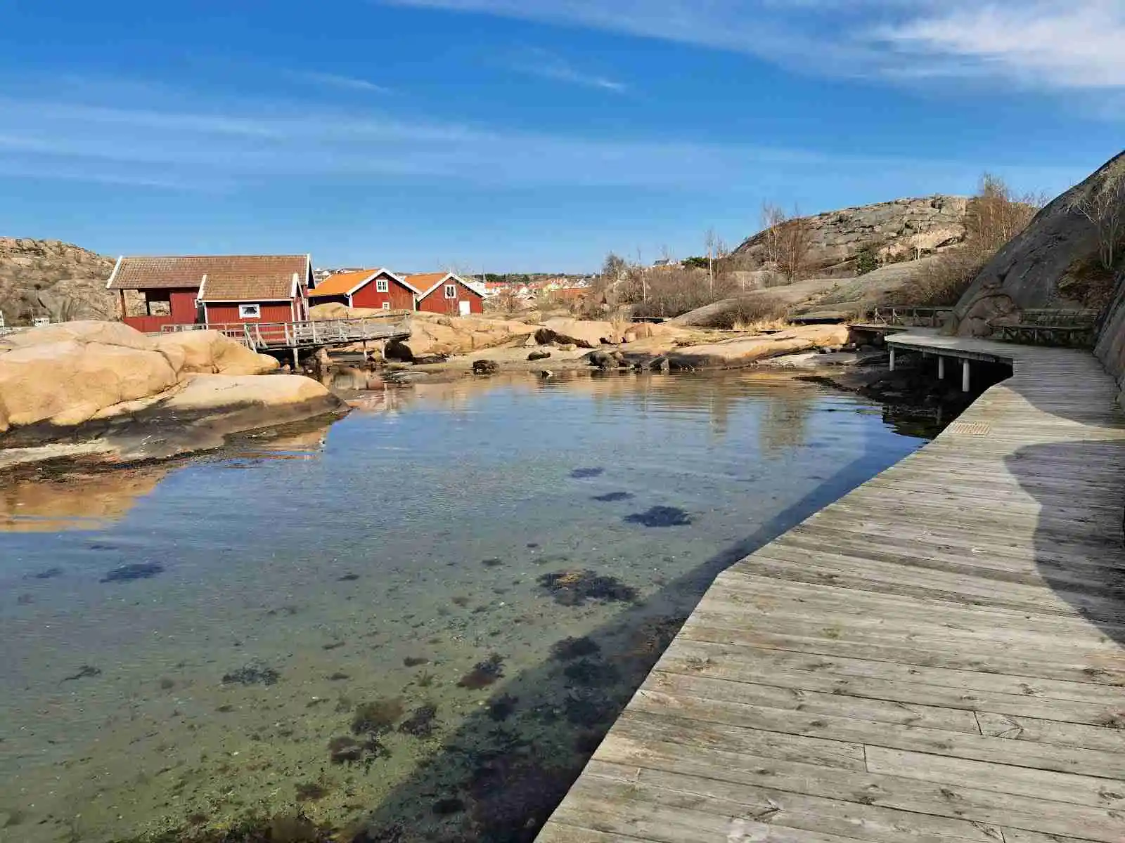Bild som visar en naturskön kustlinje vid Hunnebostrands camping och stugor, med en träbrygga som sträcker sig ut mot ett stilla vatten. Omgivande landskap inkluderar små vikar och klippiga uddar typiska för kust- och oceaniska landformer.