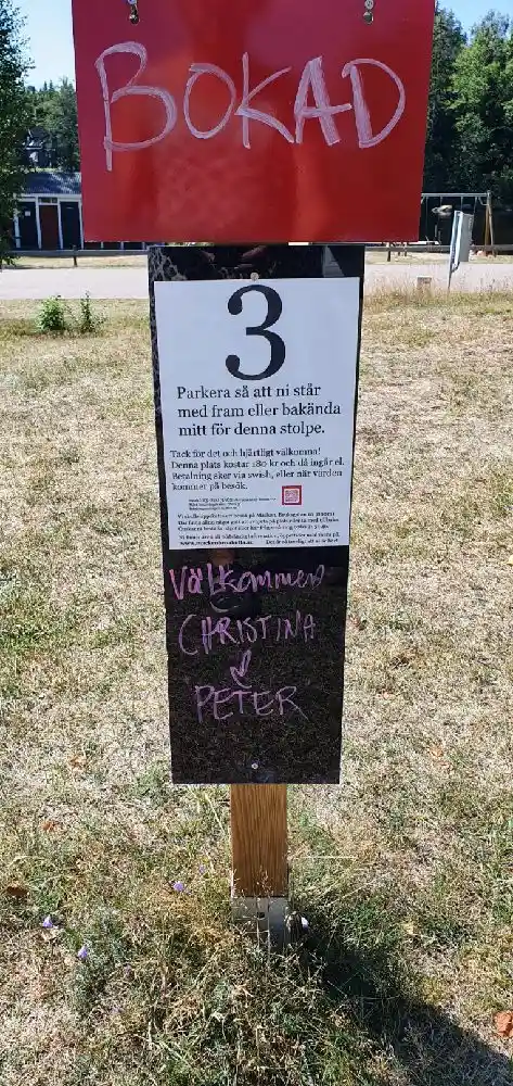 En skylt vid Broakulla Ställplats & Camping, omgiven av jord och gröna växter, med reklam och handskriven text som välkomnar besökare.