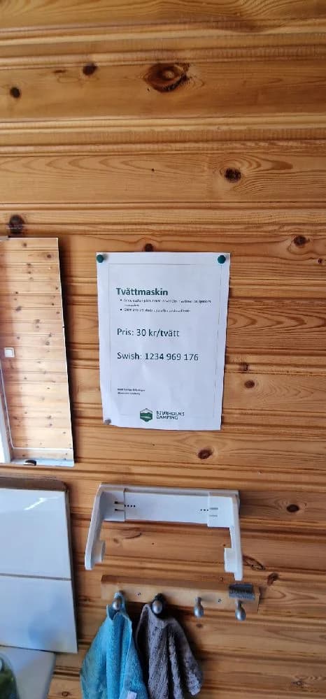 En trästuga med mörk träpanel och en veranda av trä står omgiven av grönska på Bjurholms camping. Stugan ger en hemtrevlig och rustik känsla, med väl underhållna trädetaljer som smälter in i den naturnära miljön.