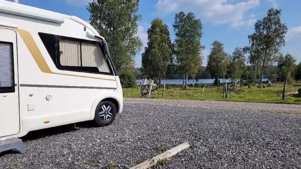 En vit husvagn parkerad på en grön campingplats, omgiven av träd och gräs, under en klarblå himmel vid Stjernkvist’s Hunshult.