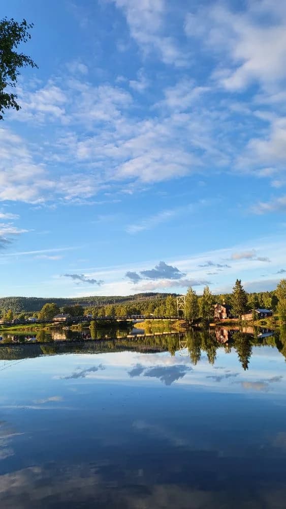 En klarblå himmel med vita moln speglas i det lugna vattnet vid Granö Beckasin, omgiven av grönska.