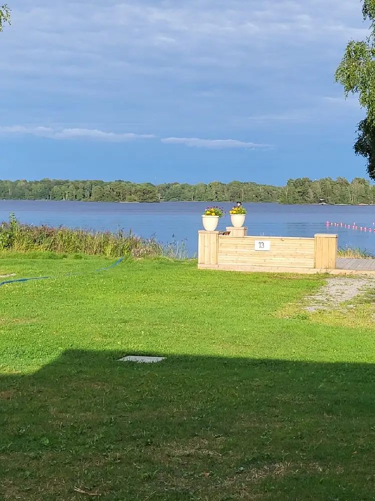 Bild av Öjaby Camping med en vidsträckt grön gräsmatta under en klarblå sommarsky. Vita moln svävar över det öppna landskapet.