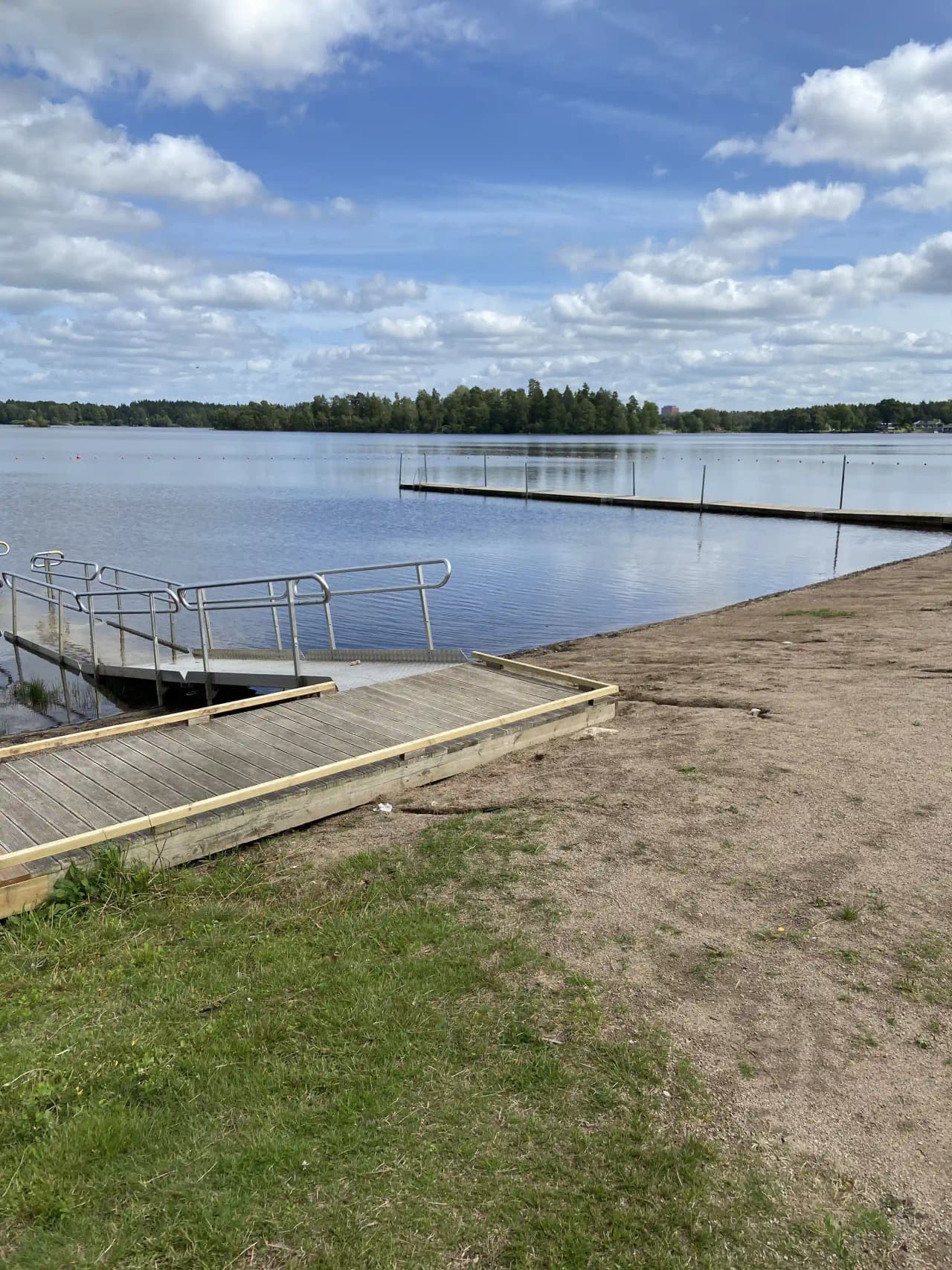 En solig dag vid Öjaby camping med moln på himlen, en brygga leder ner till en lugn sjö omgiven av grönska.